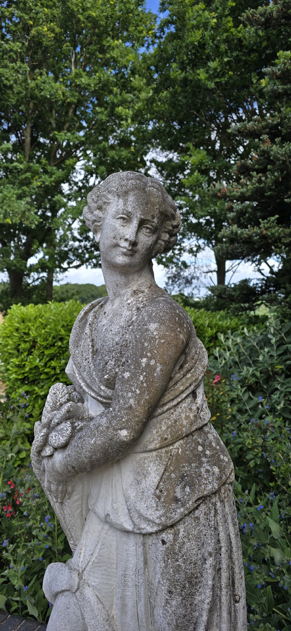 A full-length cast stone statue of a classical draped female figure standing atop a square carved plinth base. View VI