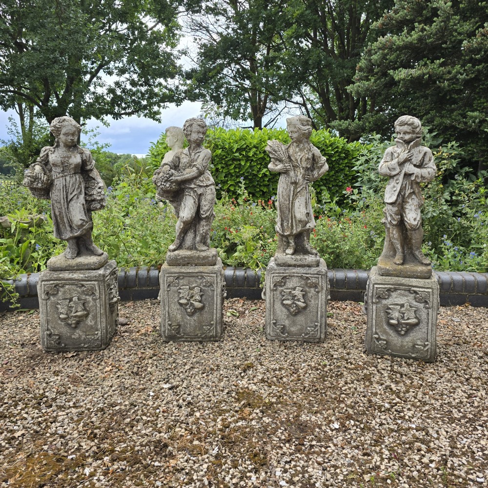 A set of four weathered grey cast stone statues representing the Four Seasons standing on matching square relief-decorated pedestals.