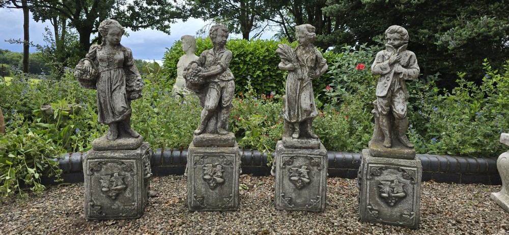 A set of four weathered grey cast stone statues representing the Four Seasons standing on matching square relief-decorated pedestals. View II