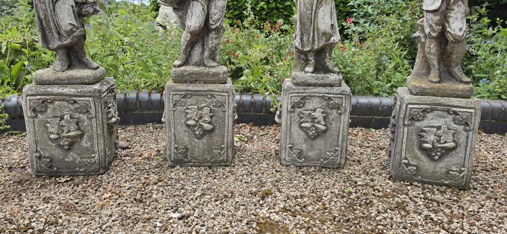 A set of four weathered grey cast stone statues representing the Four Seasons standing on matching square relief-decorated pedestals. View III