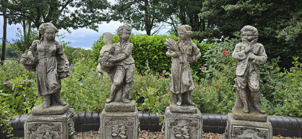 A set of four weathered grey cast stone statues representing the Four Seasons standing on matching square relief-decorated pedestals. View IV