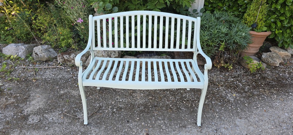 A pale green painted metal garden bench featuring a vertical slatted arched backrest, horizontal slatted seat, and curved armrests.