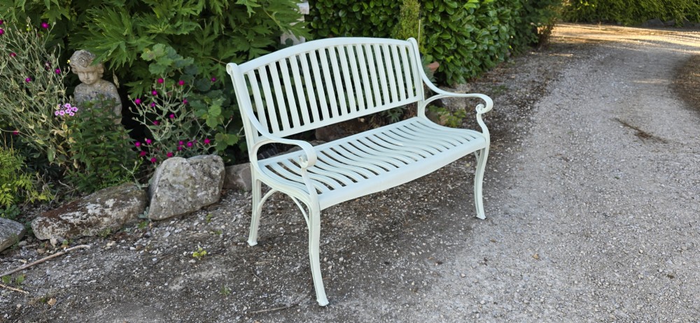 A pale green painted metal garden bench featuring a vertical slatted arched backrest, horizontal slatted seat, and curved armrests. View IV