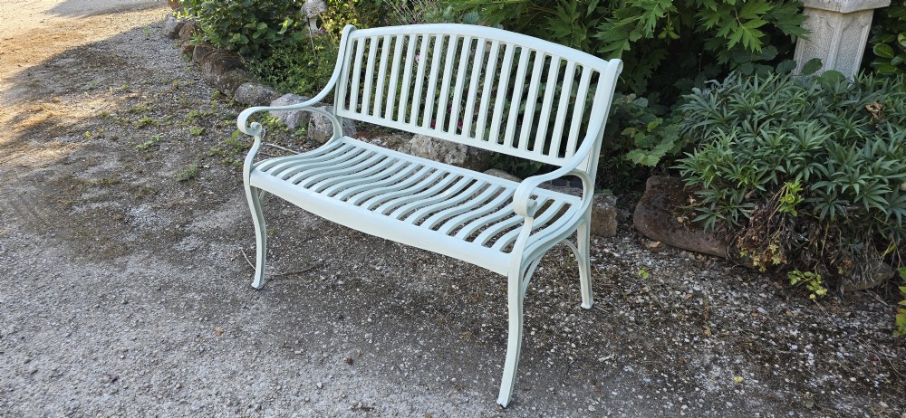A pale green painted metal garden bench featuring a vertical slatted arched backrest, horizontal slatted seat, and curved armrests. View VI