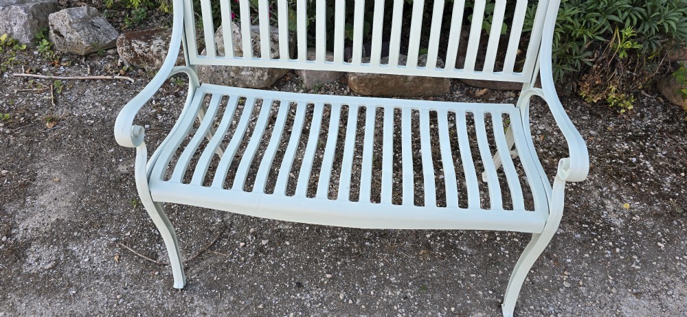 A pale green painted metal garden bench featuring a vertical slatted arched backrest, horizontal slatted seat, and curved armrests. View VII