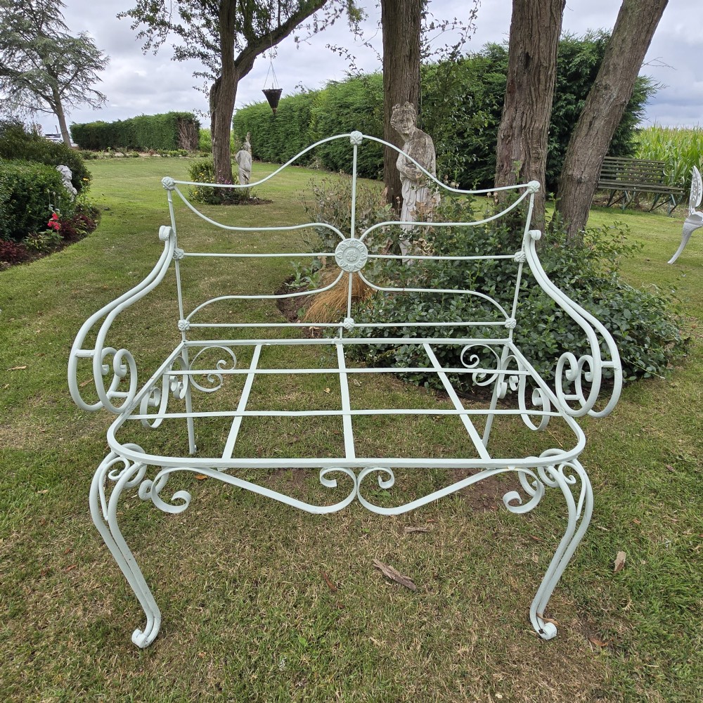 A sage green painted wrought iron garden bench featuring scrolled armrests, an arched backrest with a central rosette medallion, and an open metal grid seat.