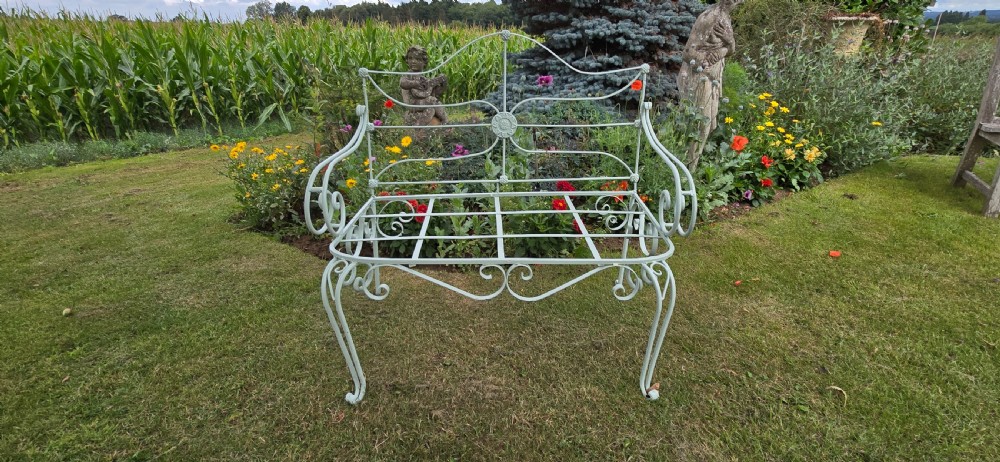 A sage green painted wrought iron garden bench featuring scrolled armrests, an arched backrest with a central rosette medallion, and an open metal grid seat. View III
