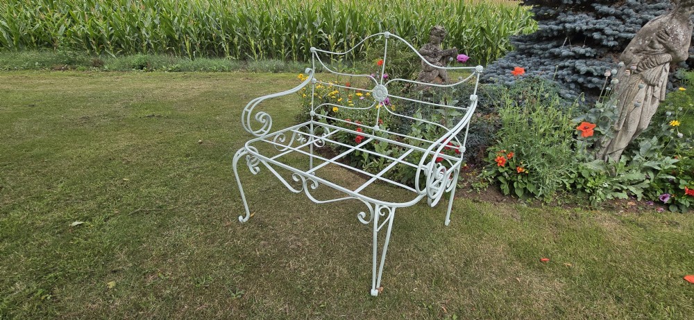 A sage green painted wrought iron garden bench featuring scrolled armrests, an arched backrest with a central rosette medallion, and an open metal grid seat. View V