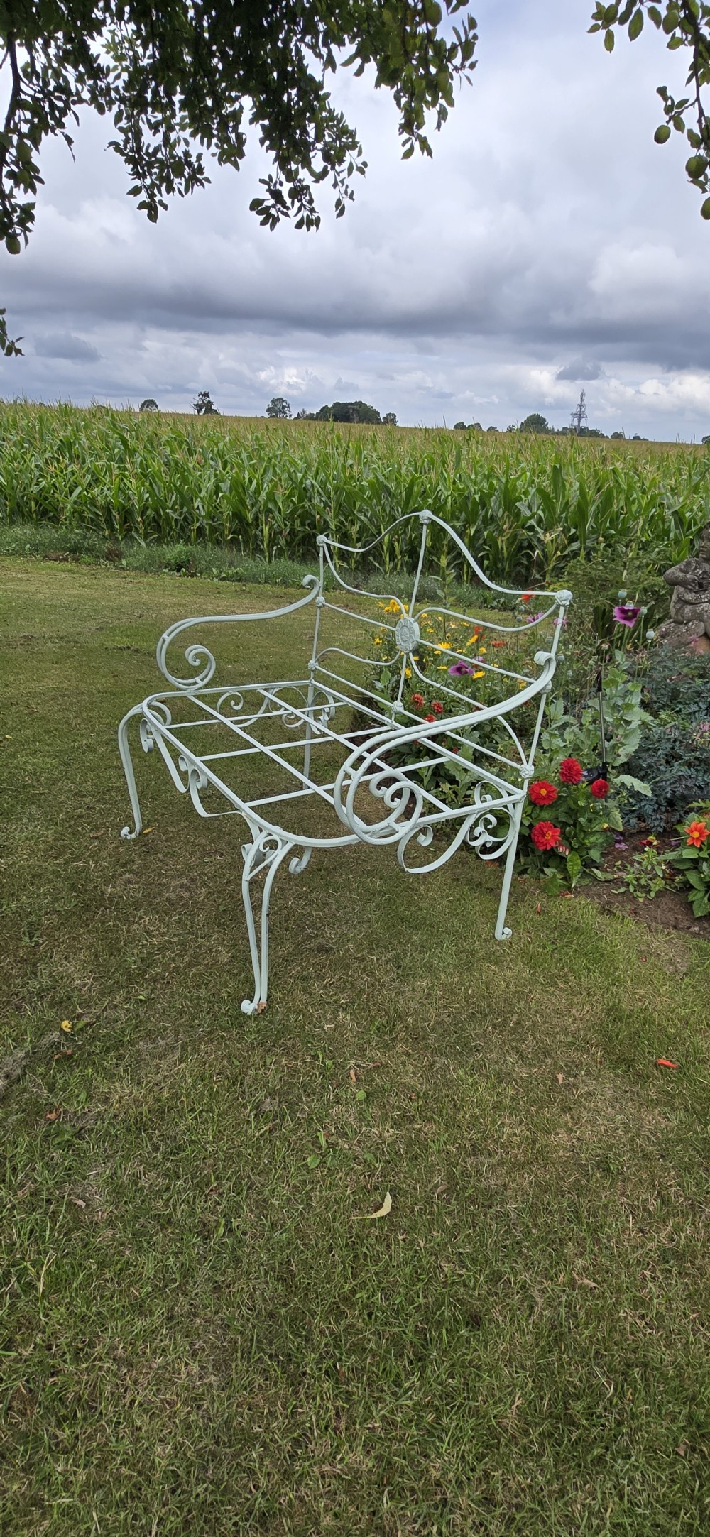 A sage green painted wrought iron garden bench featuring scrolled armrests, an arched backrest with a central rosette medallion, and an open metal grid seat. View VI