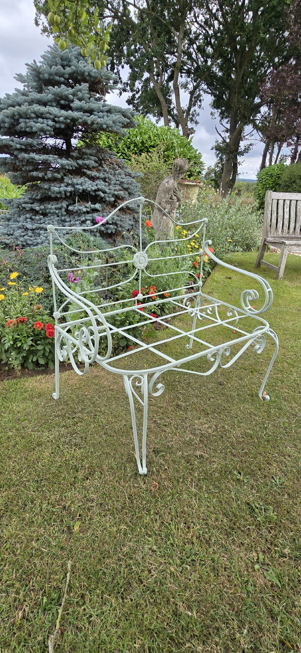 A sage green painted wrought iron garden bench featuring scrolled armrests, an arched backrest with a central rosette medallion, and an open metal grid seat. View VII