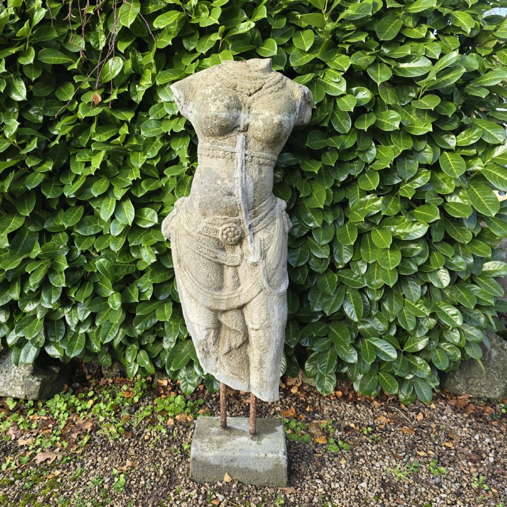 A weathered grey carved stone female torso displaying sculpted drapery and neck ornaments, elevated on two iron rods atop a square base.
