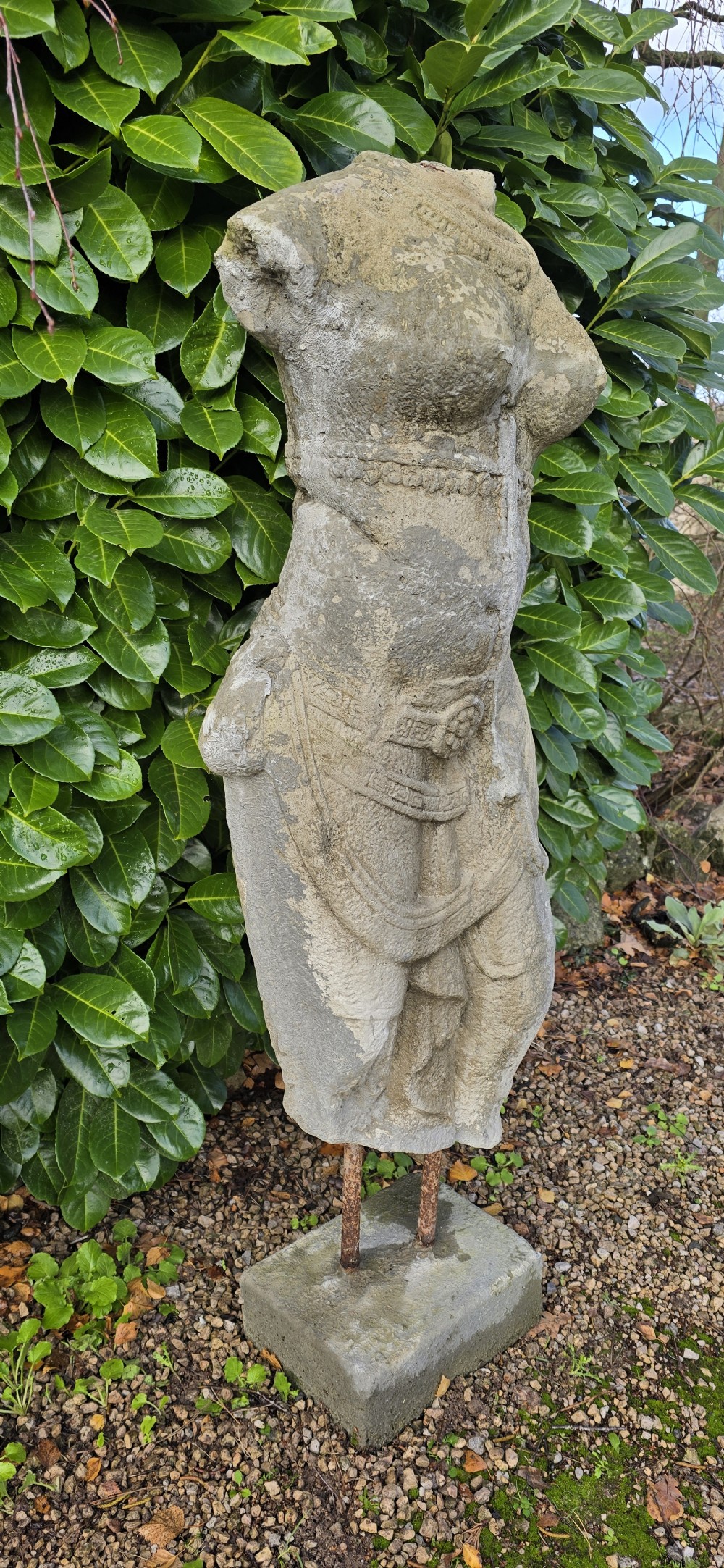 A weathered grey carved stone female torso displaying sculpted drapery and neck ornaments, elevated on two iron rods atop a square base. View III