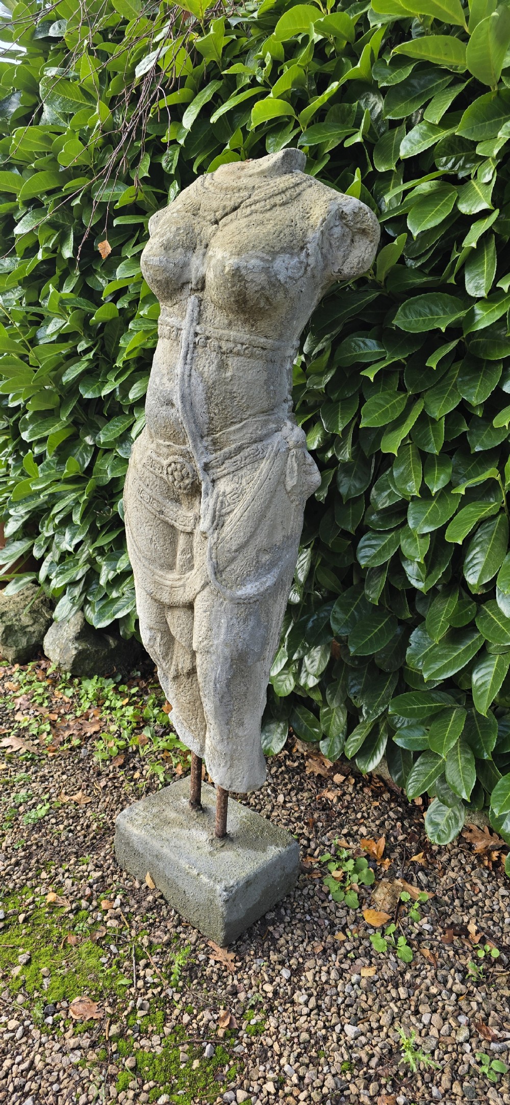 A weathered grey carved stone female torso displaying sculpted drapery and neck ornaments, elevated on two iron rods atop a square base. View IV