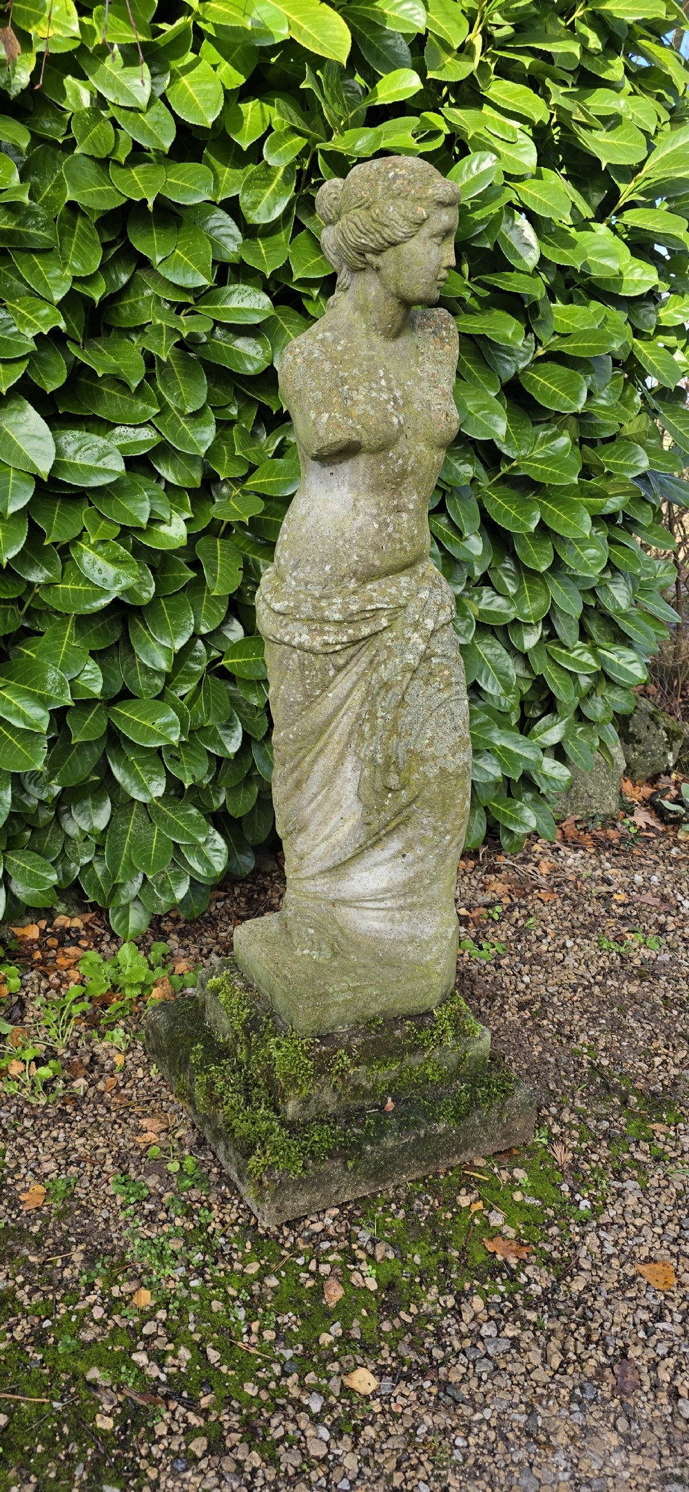 A weathered cast stone statue depicting the armless Venus de Milo draped in cloth on a square plinth. View II