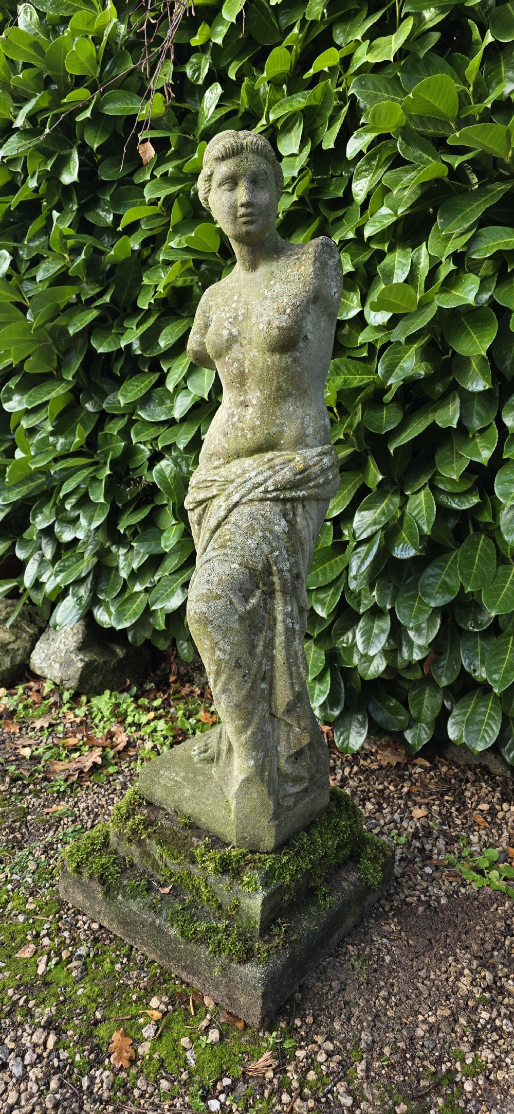 A weathered cast stone statue depicting the armless Venus de Milo draped in cloth on a square plinth. View III