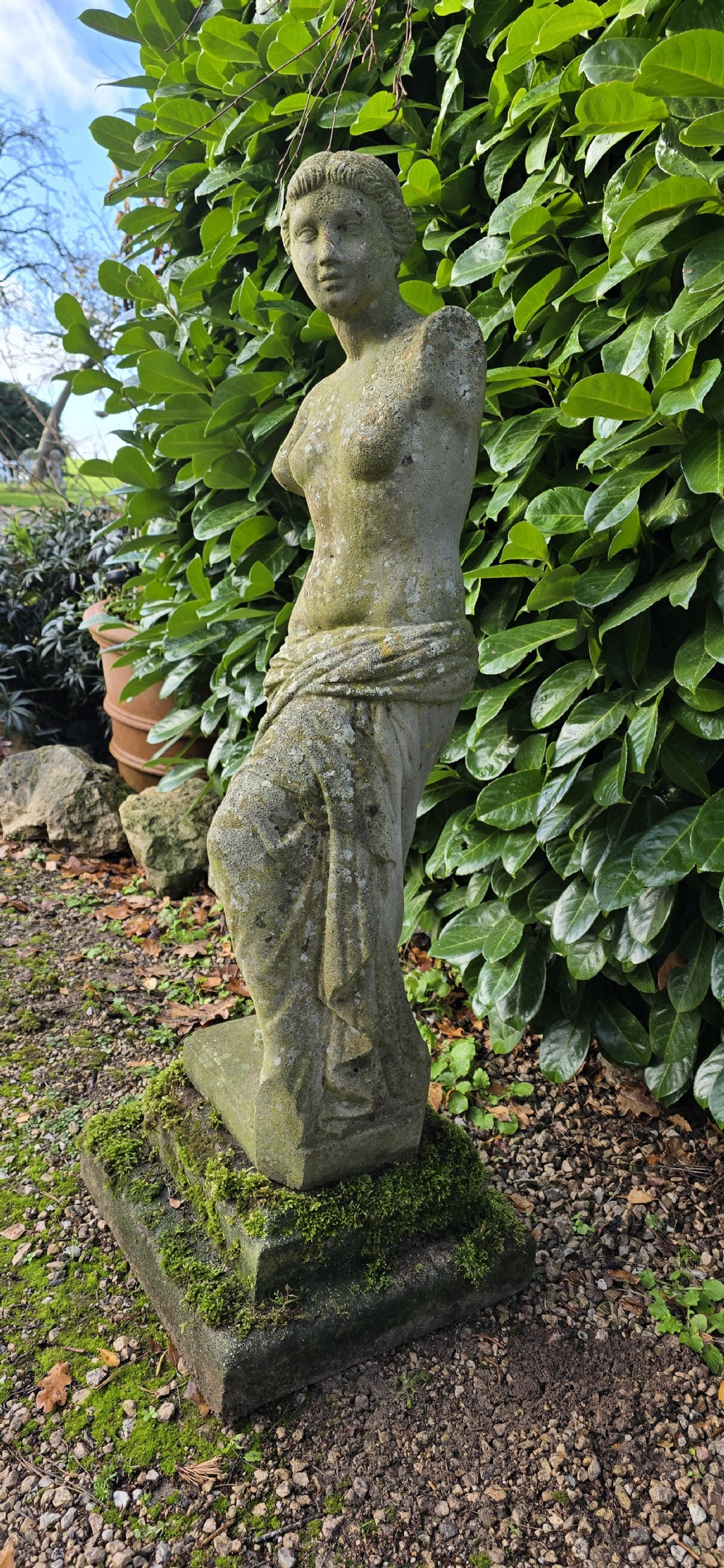 A weathered cast stone statue depicting the armless Venus de Milo draped in cloth on a square plinth. View IV