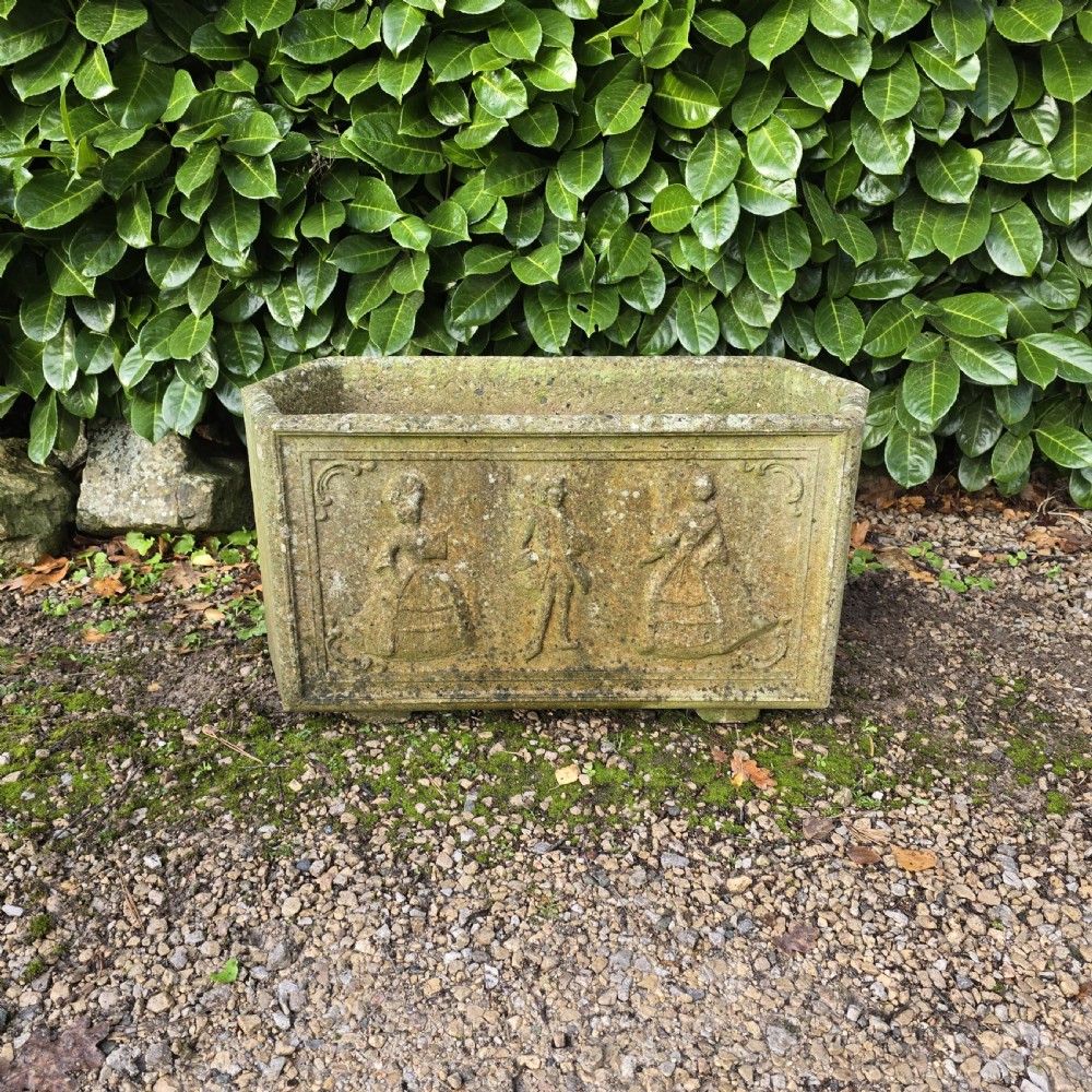 A rectangular composite stone garden trough featuring raised relief carvings of three figures in 18th-century dress on the front panel with a weathered surface.