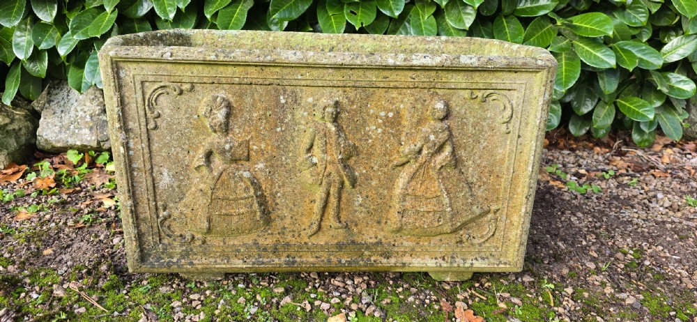 A rectangular composite stone garden trough featuring raised relief carvings of three figures in 18th-century dress on the front panel with a weathered surface. View II