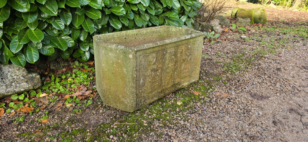 A rectangular composite stone garden trough featuring raised relief carvings of three figures in 18th-century dress on the front panel with a weathered surface. View VI