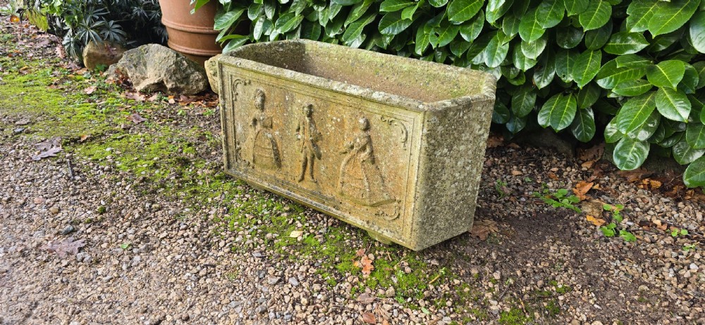 A rectangular composite stone garden trough featuring raised relief carvings of three figures in 18th-century dress on the front panel with a weathered surface. View VII