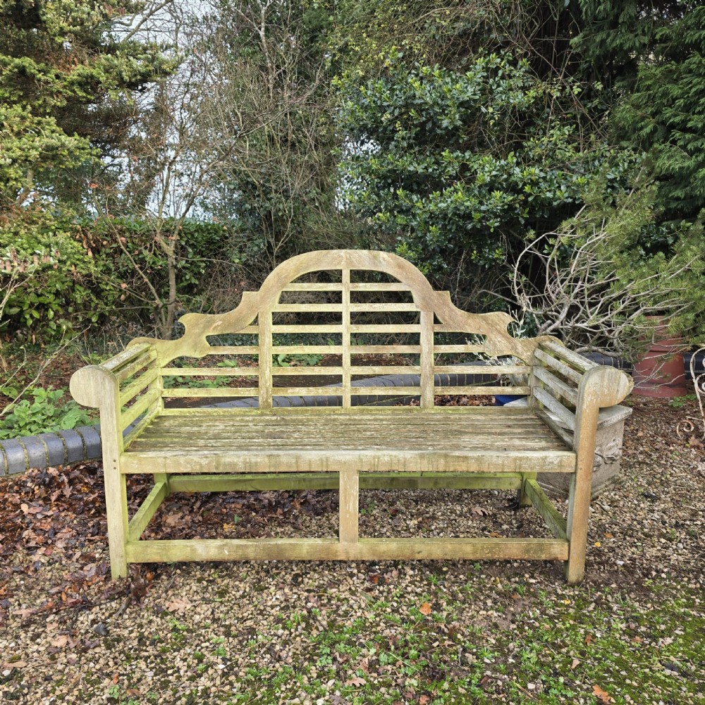 A weathered silvered teak garden bench designed in the Lutyens style with an arched slatted backrest and heavy scrolled arms.