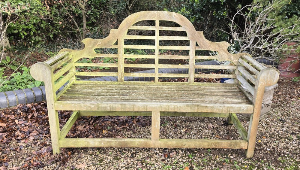 A weathered silvered teak garden bench designed in the Lutyens style with an arched slatted backrest and heavy scrolled arms. View II