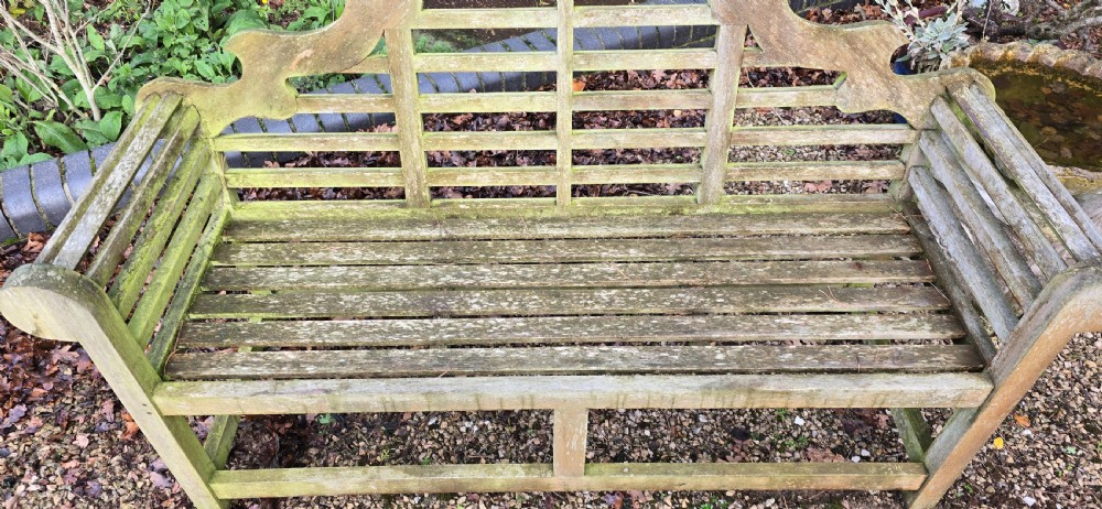 A weathered silvered teak garden bench designed in the Lutyens style with an arched slatted backrest and heavy scrolled arms. View III