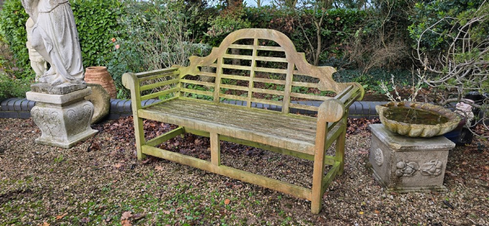 A weathered silvered teak garden bench designed in the Lutyens style with an arched slatted backrest and heavy scrolled arms. View V