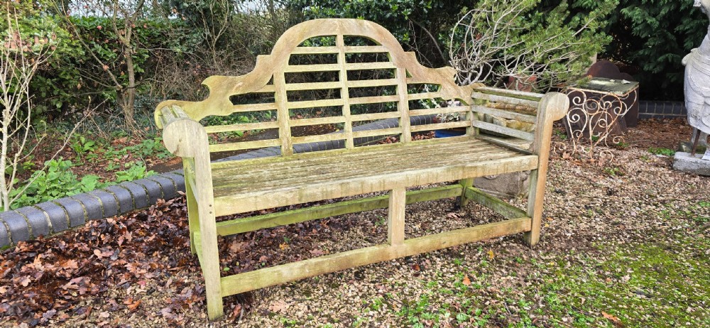 A weathered silvered teak garden bench designed in the Lutyens style with an arched slatted backrest and heavy scrolled arms. View VI