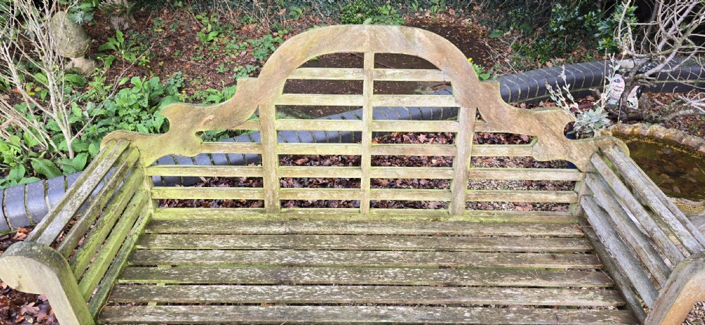 A weathered silvered teak garden bench designed in the Lutyens style with an arched slatted backrest and heavy scrolled arms. View VII