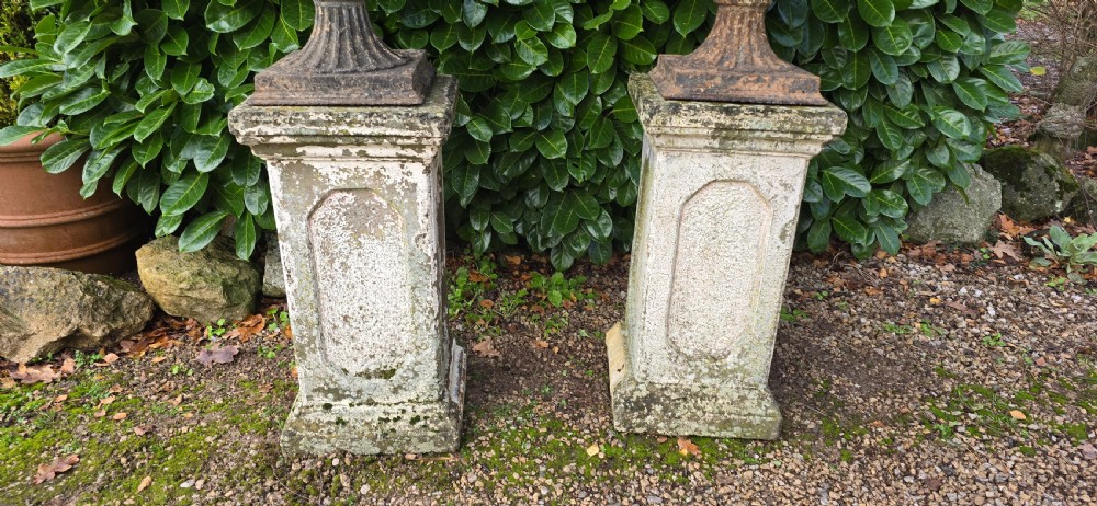 A pair of weathered cast iron garden urns with fluted rims resting on tall, square stone plinths. View II