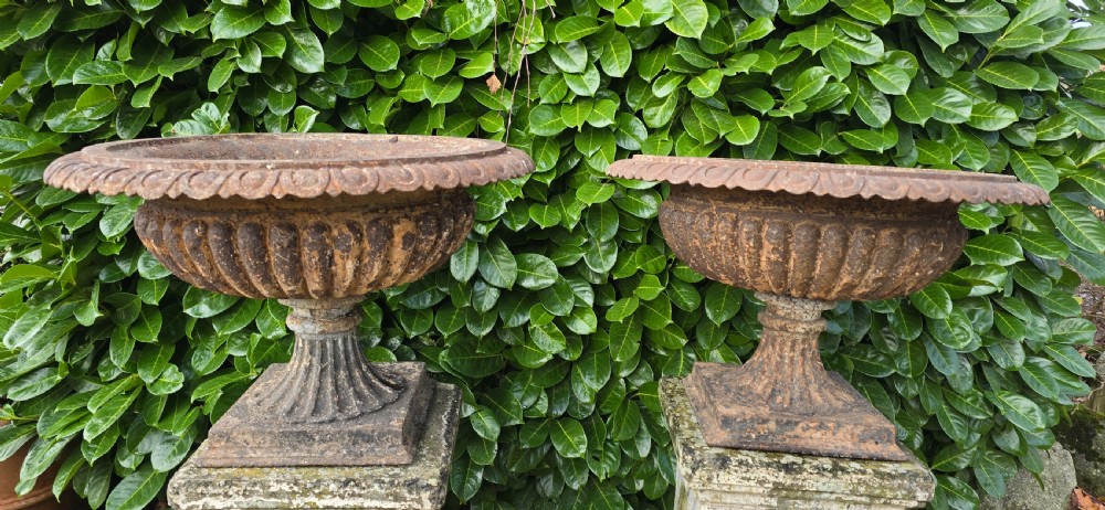 A pair of weathered cast iron garden urns with fluted rims resting on tall, square stone plinths. View III