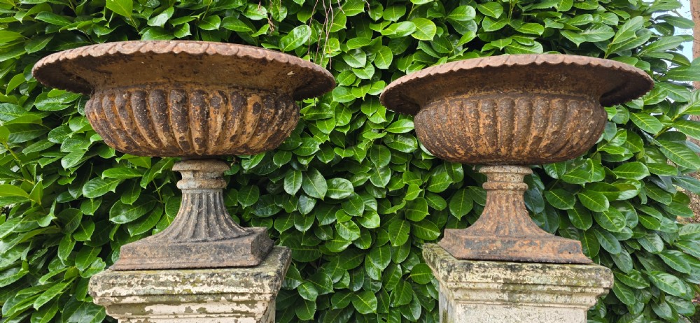 A pair of weathered cast iron garden urns with fluted rims resting on tall, square stone plinths. View V