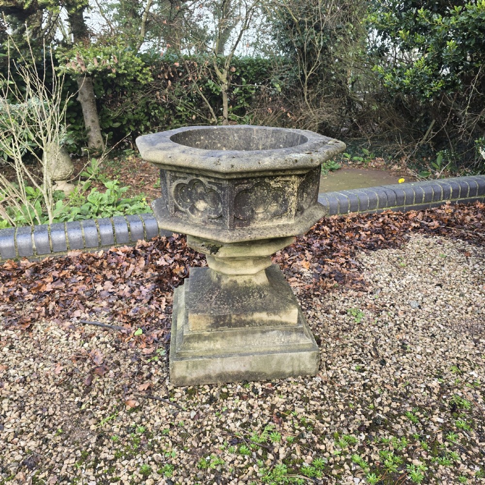 A weathered heptagonal cast stone garden urn featuring carved Gothic tracery panels resting on a stepped pedestal base.