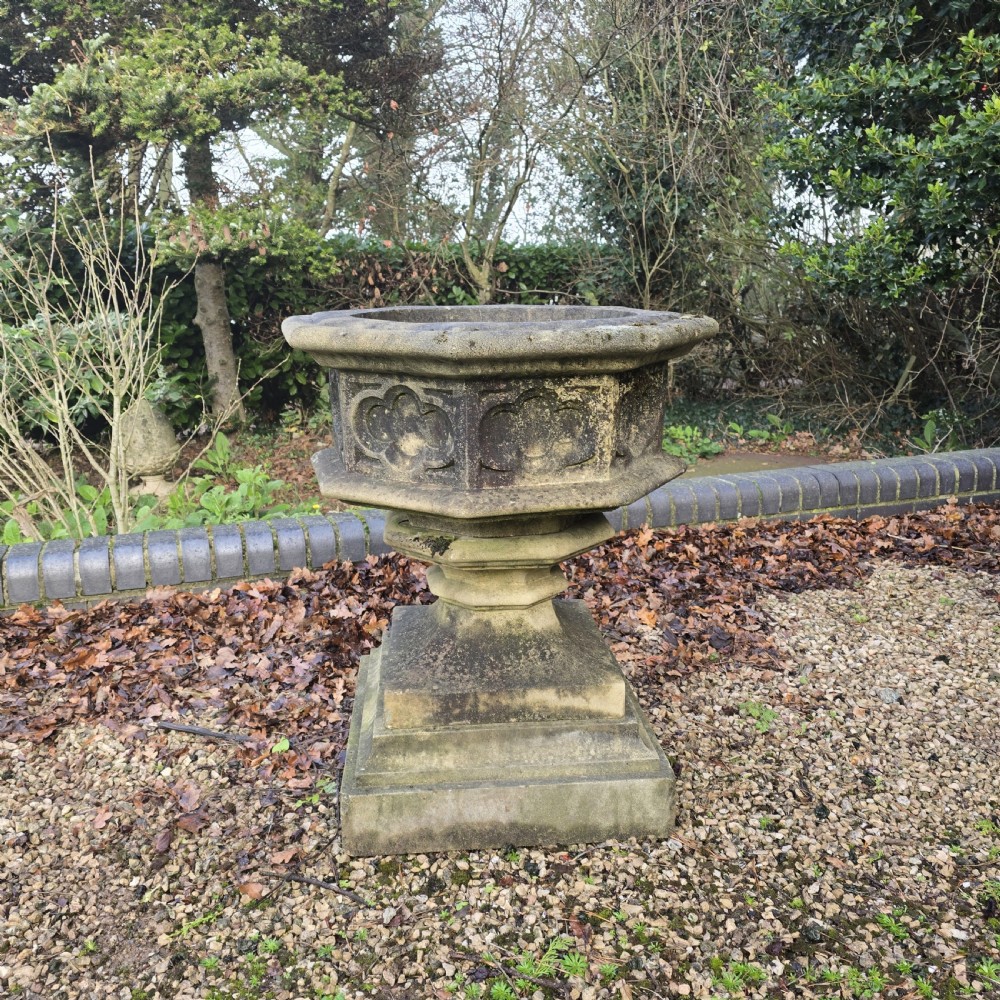 A weathered heptagonal cast stone garden urn featuring carved Gothic tracery panels resting on a stepped pedestal base. View II