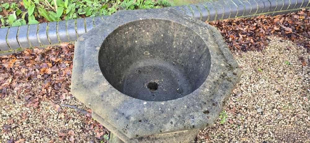 A weathered heptagonal cast stone garden urn featuring carved Gothic tracery panels resting on a stepped pedestal base. View V
