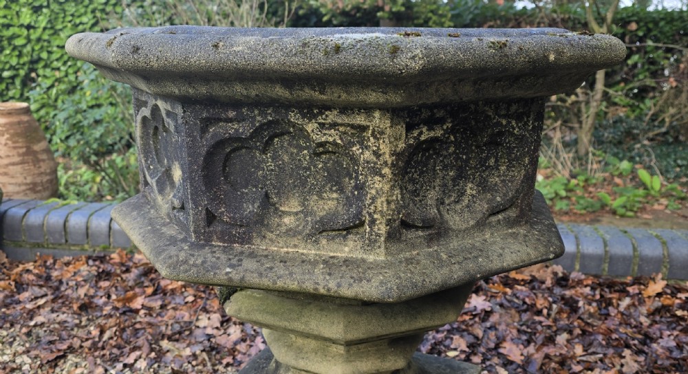A weathered heptagonal cast stone garden urn featuring carved Gothic tracery panels resting on a stepped pedestal base. View VII