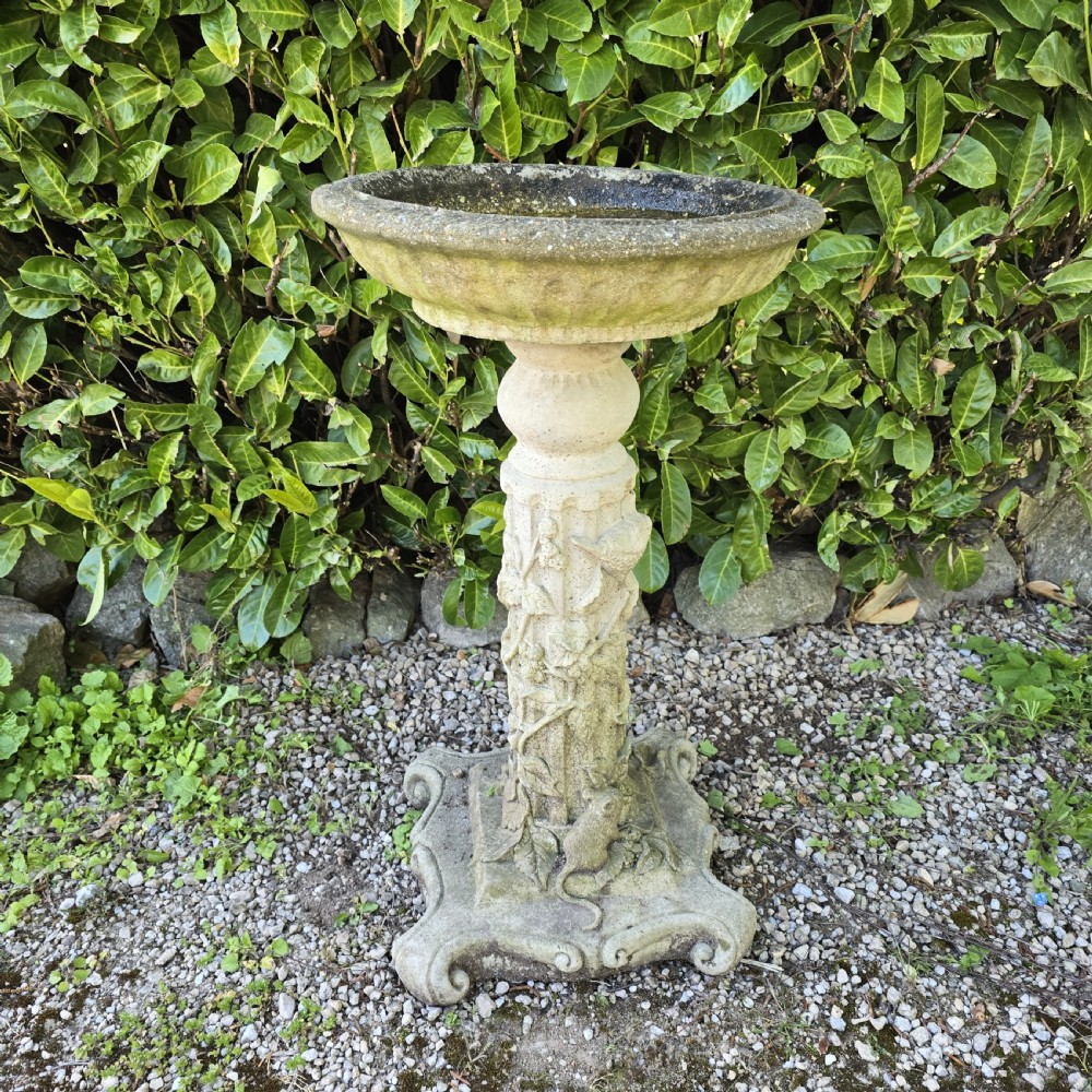 A weathered cast stone pedestal bird bath featuring relief carvings of foliage, birds, and a mouse around the column, resting on a scroll-cornered base. View II