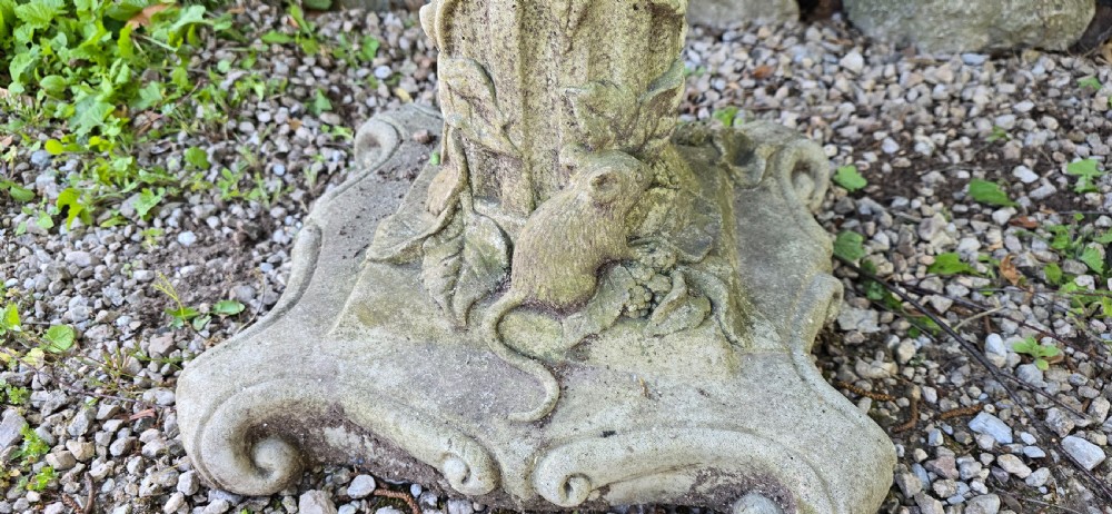 A weathered cast stone pedestal bird bath featuring relief carvings of foliage, birds, and a mouse around the column, resting on a scroll-cornered base. View VI