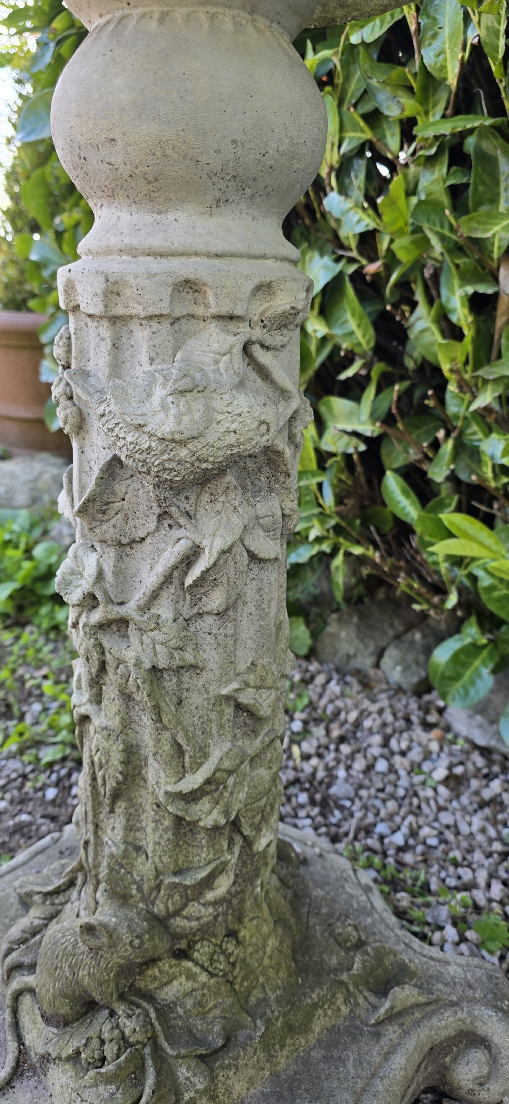 A weathered cast stone pedestal bird bath featuring relief carvings of foliage, birds, and a mouse around the column, resting on a scroll-cornered base. View VII