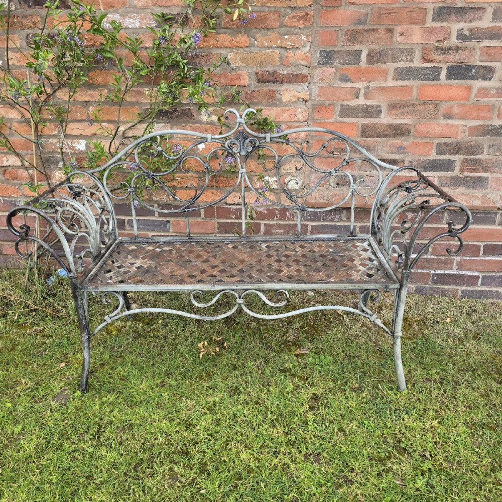 A French metal garden bench featuring a weathered grey-green painted finish, decorative scrollwork backrest, rolled arms, and a lattice seat.