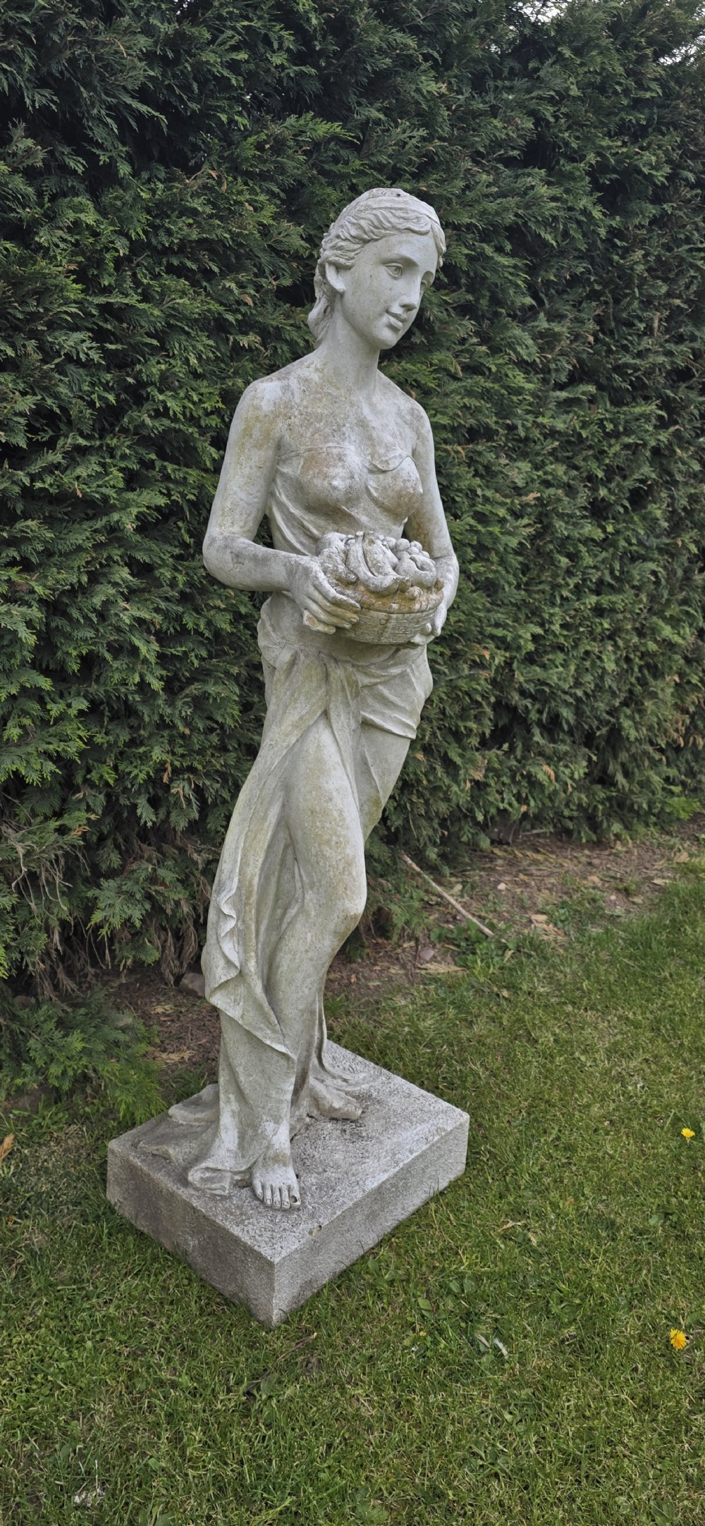 A life-sized classical stone garden statue of a draped female figure holding a basket of fruit on a square base. View II