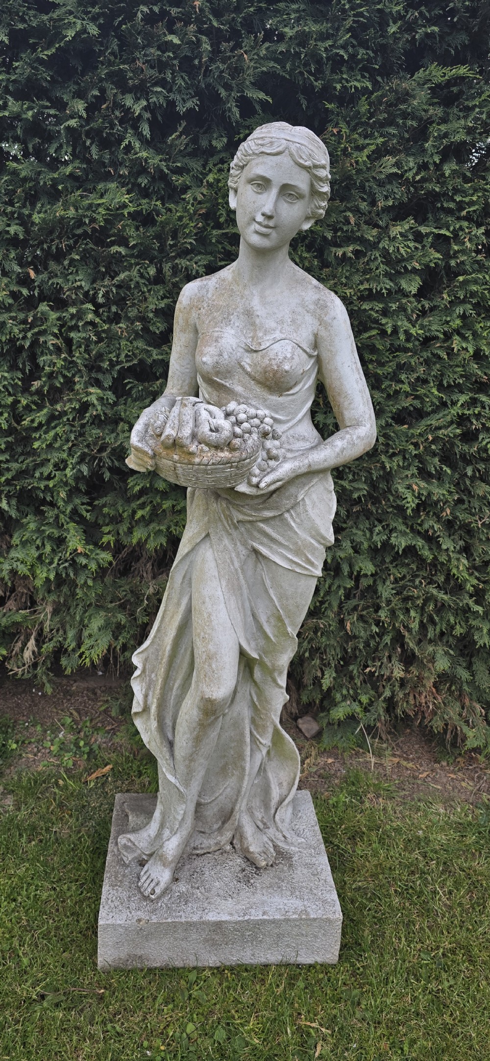 A life-sized classical stone garden statue of a draped female figure holding a basket of fruit on a square base. View III