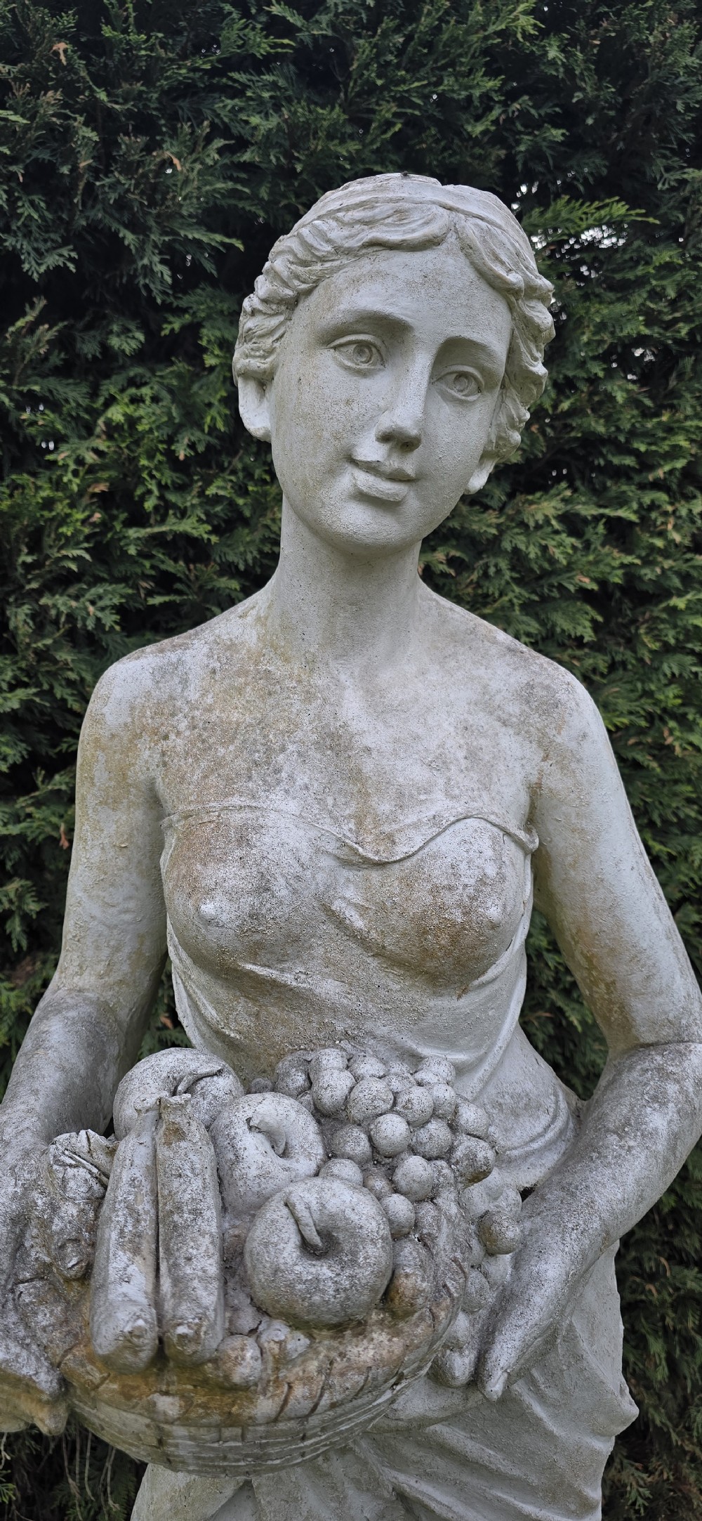A life-sized classical stone garden statue of a draped female figure holding a basket of fruit on a square base. View IV