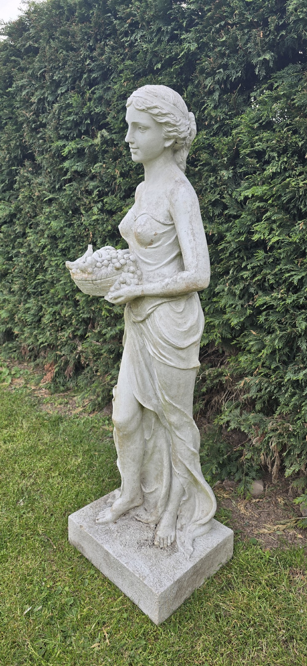 A life-sized classical stone garden statue of a draped female figure holding a basket of fruit on a square base. View VI