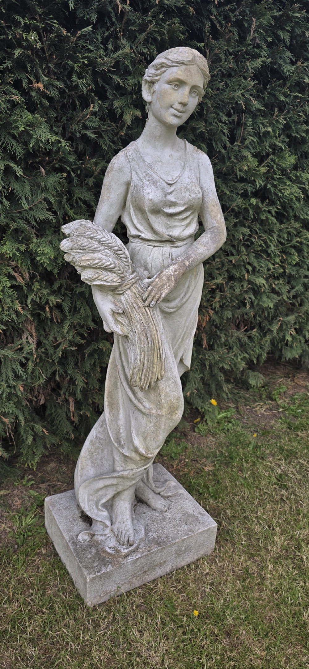 A life-sized weathered stone classical garden statue representing summer, featuring a draped female figure holding a sheaf of wheat on a square plinth base. View II