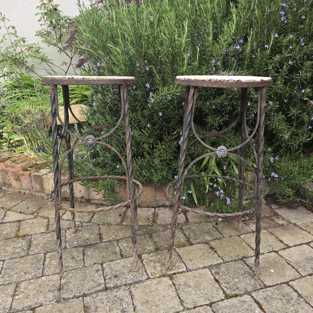 A pair of tall wrought iron plant stands with circular stone tops, twisted legs, and central lion mask medallion details.