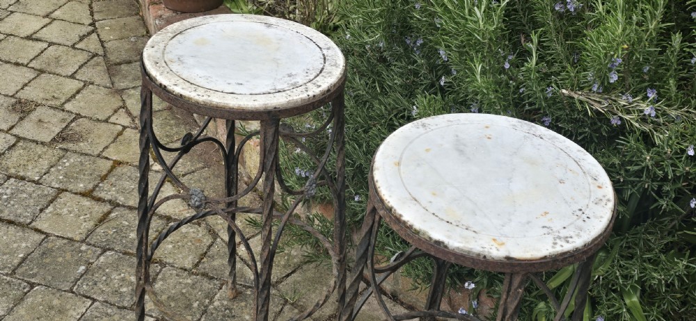 A pair of tall wrought iron plant stands with circular stone tops, twisted legs, and central lion mask medallion details. View X