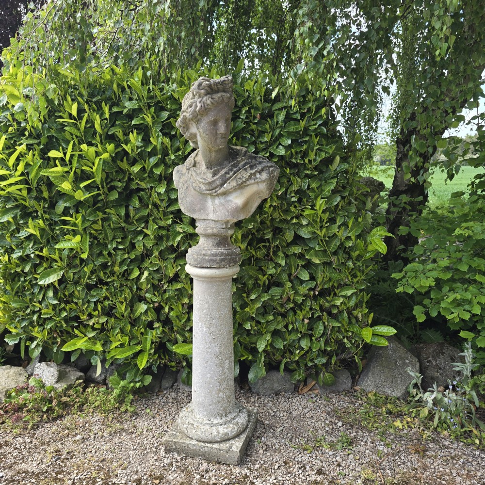 A weathered stone bust of Apollo set atop a cylindrical column pedestal with a square base.