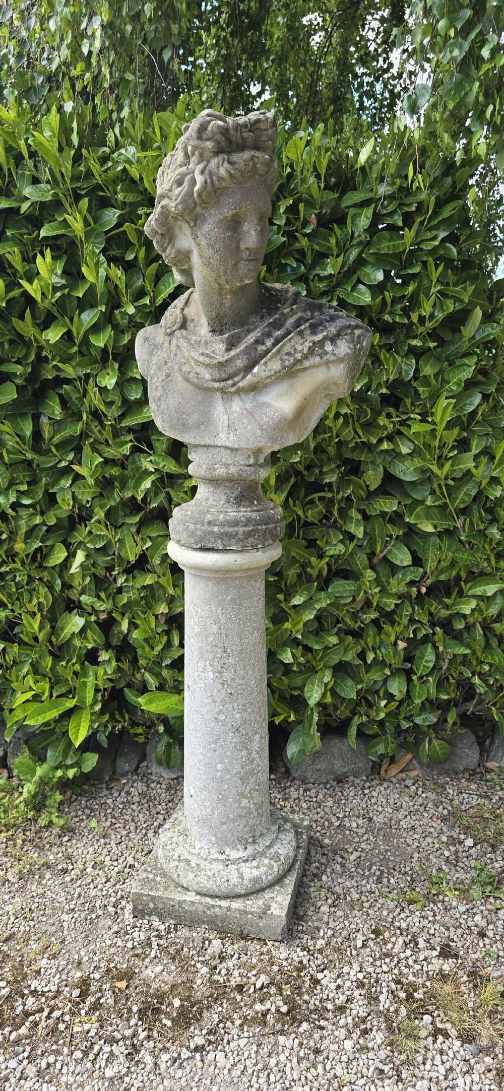 A weathered stone bust of Apollo set atop a cylindrical column pedestal with a square base. View II